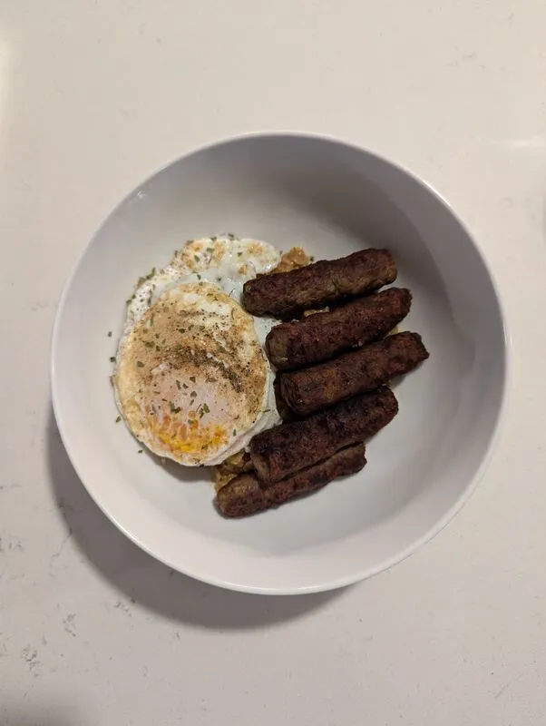 Fried eggs and sausage links in a white bowl over hash browns